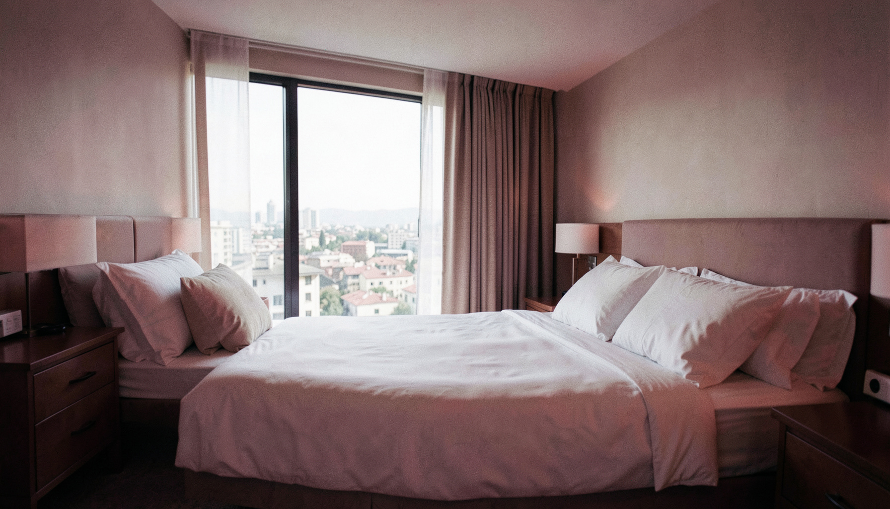 Chambre d'hôtel lumineuse avec grand lit, fenêtre offrant une vue sur la ville, tons neutres et ambiance sereine